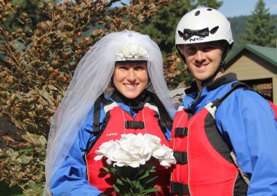 A bride and groom posing together in their rafting gear. The groom is wearing a special white rafting helmet with a bow tie and the bride is wearing a white rafting helmet with a veil on it.